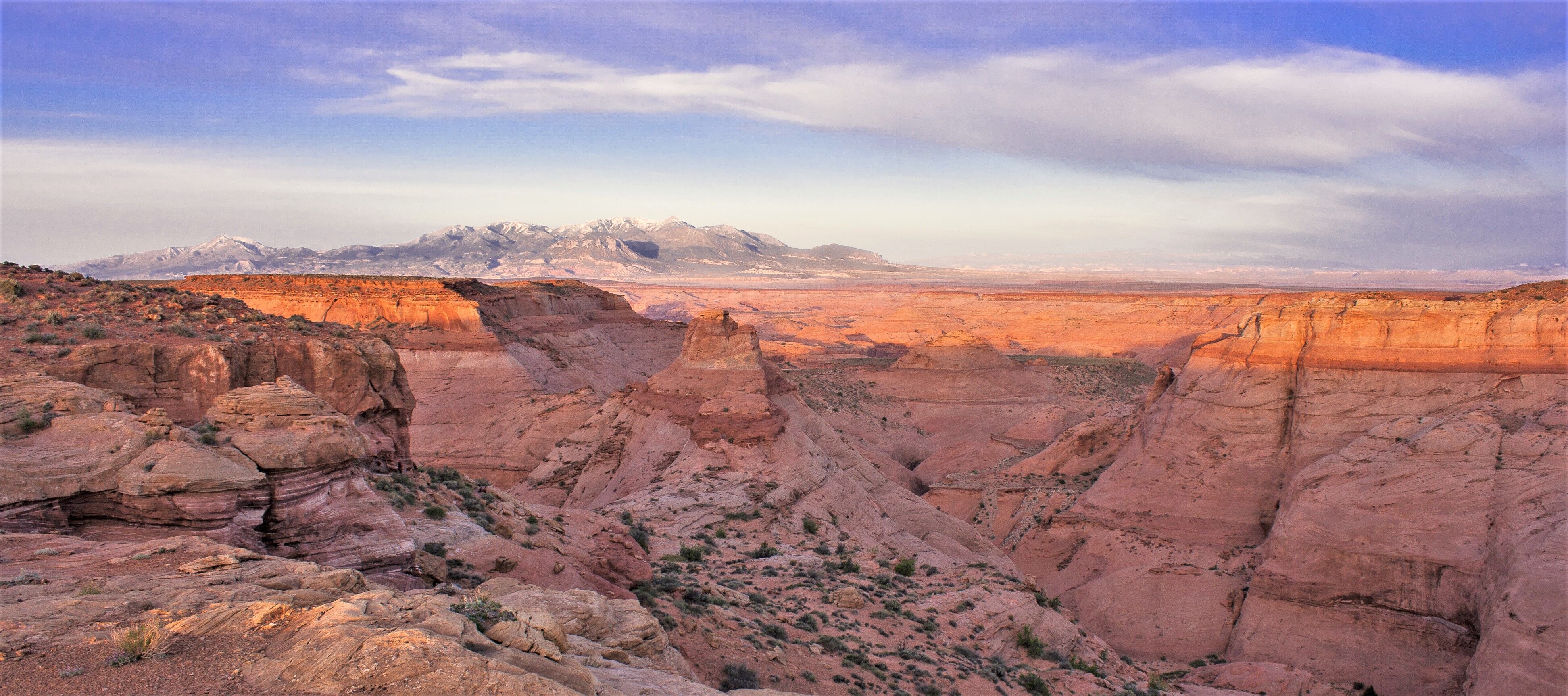 The image shows a vast canyon landscape under a blue sky with scattered clouds. The canyon walls are layered with reddish and orange hues, suggesting sedimentary rock formations. In the distance, a mountain range is visible. The foreground features rocky terrain with sparse vegetation, highlighting the arid environment. The overall scene conveys a sense of depth and natural beauty.